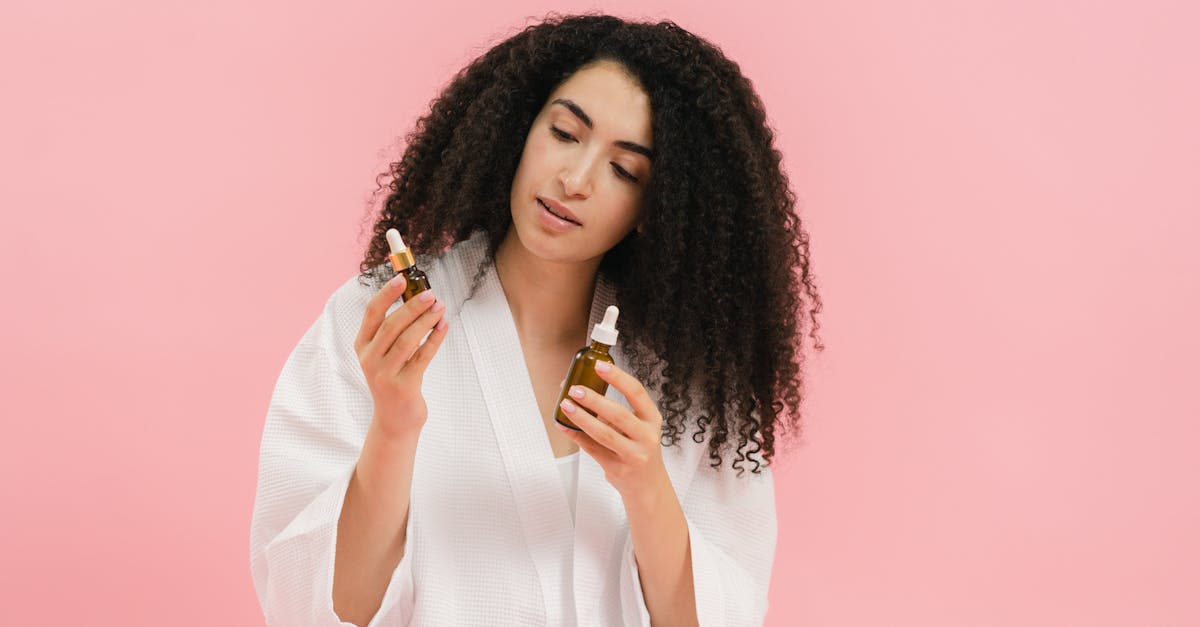 a woman with curly hair in a white robe examines two cosmetic oil bottles against a pink background